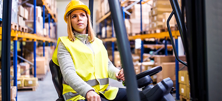 Worker driving a forklift on a loading dock with racks of inventory and products behind her.